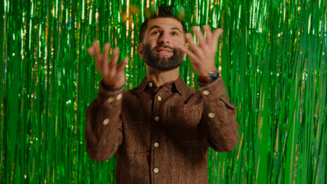 Man-Celebrating-St-Patrick's-Day-Standing-In-Front-Of-Green-Tinsel-Curtain-And-Throwing-Novelty-Gold-Coins-In-The-Air